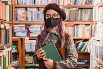 Young caucasian clever hipster high school girl pupil college student teenager wearing medical mask to protect from virus holding a book in the university library or bookshop. Education concept