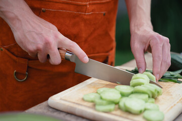 Close up of man holding sharp knife, chopping cucumber. Preparing vegetables for an outdoor barbecue.