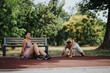 © qunica.com - Two women taking a break post-exercise in a park on a sunny day, one sitting on a bench and the other catching breath on the ground.