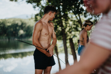 Wall Mural - Young shirtless man setting up a tent by a lake with friends in the background on a sunny day.