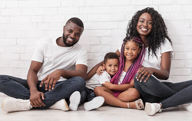 Wall Mural - Joyful afro family mother, father and two little children sitting on floor and cuddling over white brick wall background.