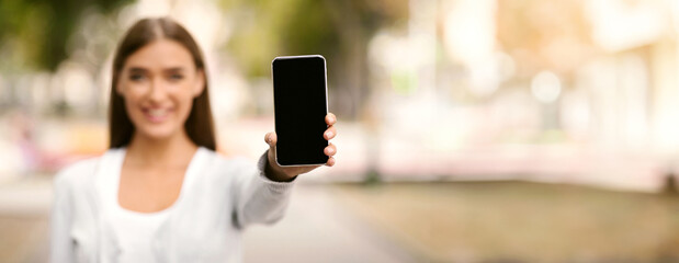 Wall Mural - Girl Showing Smartphone Empty Screen To Camera Recommending App Standing In Park Outdoor. Mobile Application. Shallow Depth, Selective Focus, Mockup