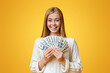 © Prostock-studio - Smart savings. Joyful young girl holding fan of dollars in hands and smiling to camera over orange studio background, copy space