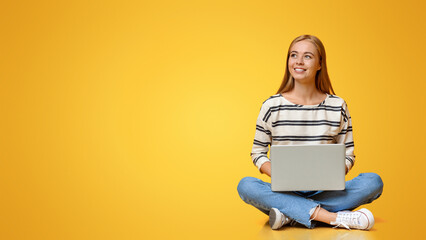 Dreamy teenage girl sitting with laptop on floor and looking aside at copy space on orange background