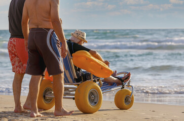 Accessible beach wheelchair helps individual enjoy the ocean at sunset. A beach wheelchair with large yellow wheels supports a person sitting in it, as two individuals stand nearby, enjoying the sea