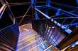 © Emvats - A detailed view of a metal staircase with grid-patterned steps, illuminated by blue and yellow lights. The intersecting lines and contrasting colors create an industrial, abstract visual.