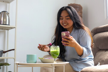 Happy Young Asian Woman Eating Healthy Food And Drink While Using Smartphone At Home