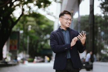 Confident Asian businessman in modern city, smiling while using smartphone outdoors, wearing professional suit, representing success and connectivity in digital age.