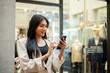 © bongkarn - A confident, happy Asian woman is reading messages on her phone in front of a clothing store.