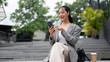 © bongkarn - A happy Asian businesswoman is replying chat on her smartphone while sitting on stairs outdoors.