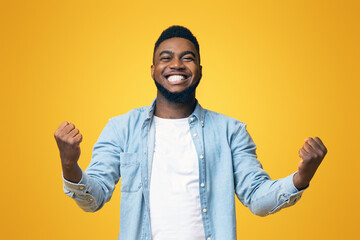 Joyful african american man rejoicing success with clenched fists on yellow studio background with copy space, panorama