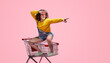 © kegfire - Cheerful woman in casual outfit, sitting in a shopping cart and pointing forward. Fun and playful concept against a bright pink background.
