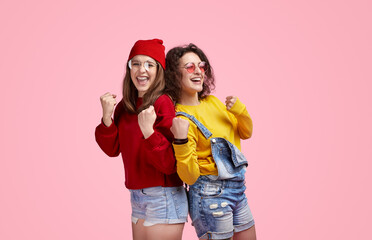 Wall Mural - Two joyful young women celebrating with excitement, wearing casual colorful clothes on a pink background. Friendship and happiness concept.