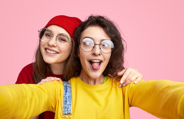 Wall Mural - Two young friends wearing glasses taking a fun selfie together, one sticking out her tongue, against a bright pink background.