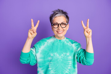 Wall Mural - Photo portrait of attractive senior woman show v-sign dressed stylish green clothes isolated on purple color background