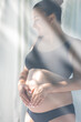 © puhimec - A pregnant woman carrying her large belly while standing by the window.