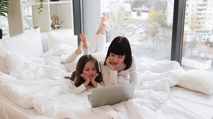 Wall Mural - Mother and daughter relaxing on bed enjoying quality time together. Both wearing comfortable clothing, using laptop, engaging in conversation. Bright natural light from window, cozy home environment.