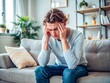© Sirinporn - Young adult suffering from vertigo or migraine pain, clutching head with hand while sitting on a couch in a living room, conveying discomfort and distress.