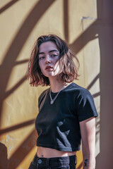 Wall Mural - Young woman with a bob haircut in a black crop top, standing against a yellow wall with dramatic shadows, looking confidently at the camera