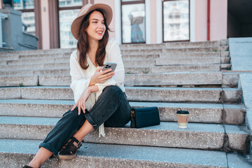 Wall Mural - Young beautiful smiling woman in trendy summer clothes. Carefree female posing in street in sunny day. Positive model holds smartphone, looks at cellphone screen, uses mobile apps, in hat