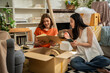 © SKW - Two women sitting on a checkered rug in a room with boxes and books