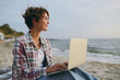 © ViDi Studio - Side view young IT woman wear shirt casual clothes hold use work on laptop pc computer look aside sits rest on sea ocean sand shore beach outdoor seaside in summer day free time. Lifestyle concept.