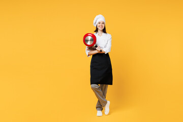 Full body young smiling happy fun chef cook baker woman wear white shirt black apron uniform toque chefs hat hold in hand frying pan isolated on plain yellow background studio. Cooking food concept.