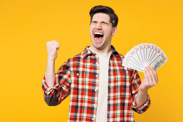 Wall Mural - Young happy man he wears red checkered shirt casual clothes hold in hand fan of cash money in dollar banknotes do winner gesture isolated on plain yellow orange background studio. Lifestyle concept.