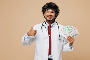 Wall Mural - Young Indian doctor man he wear white medical gown suit work in hospital clinic hold fan of cash money in dollar banknotes show thumb up isolated on plain beige background Healthcare medicine concept