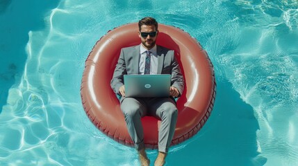 A young Caucasian man in a suit lounges on a red inflatable ring while working on a laptop in a bright blue pool.