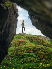 Wall Mural - View through the cave to the girl on the hill with yellow and pink flowers and green grass in alpine mountain valley at sunset in summer. Landscape with woman, grotto, rock, flowers. Dolomites, Italy