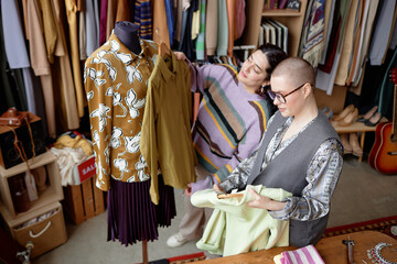 Fashionable buzz cut female stylist working with Middle Eastern client helping choose second hand outfit next to mannequin with vintage clothes at thrift store