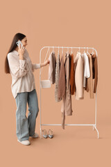 Sticker - Happy female stylist with clothes on rack talking by mobile phone against beige background