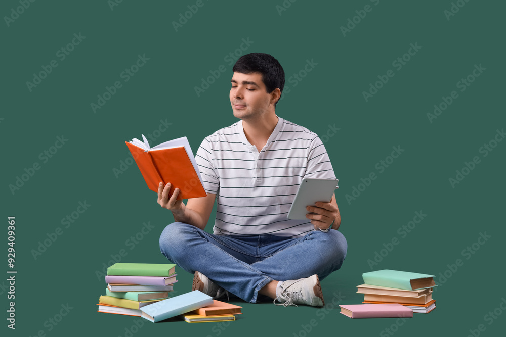 Young man with books and tablet computer sitting on green background