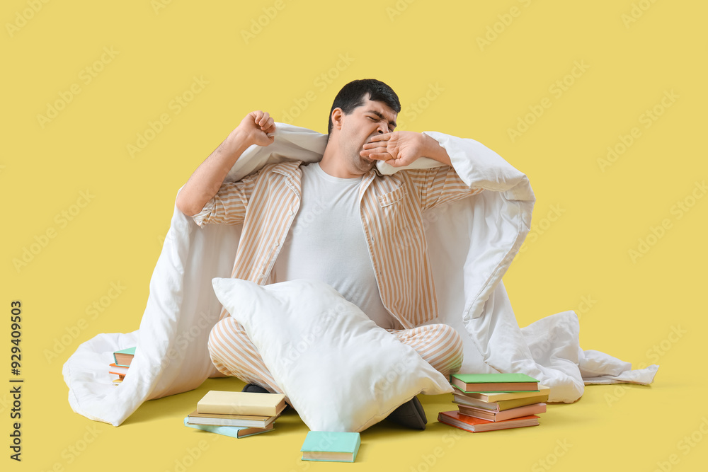 Sleepy young man with blanket and books on yellow background