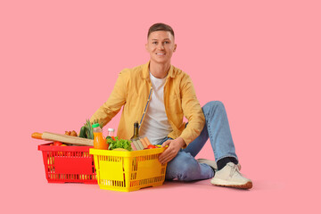 Wall Mural - Young man with full shopping baskets sitting on pink background