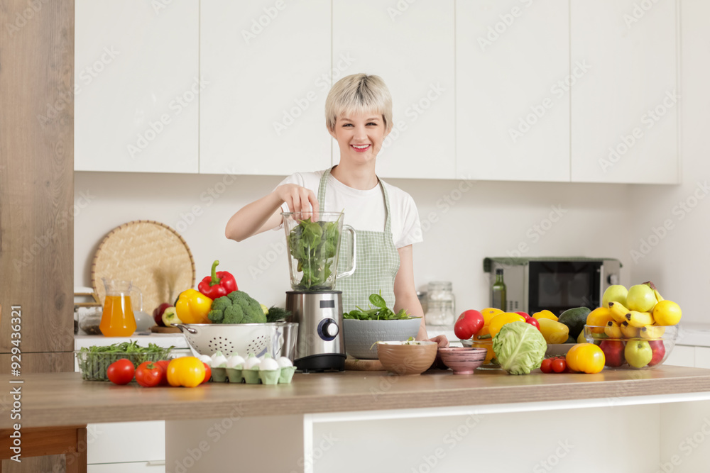 Young woman putting spinach into blender in kitchen