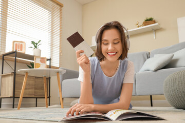 Poster - Beautiful young happy woman in headphones with tasty chocolate bar reading magazine at home