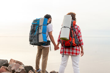 young african american couple with hiking backpacks and equipment standing against sea and sky and holding hands, curly woman and man tourists traveling looking away at horizon on isolated background