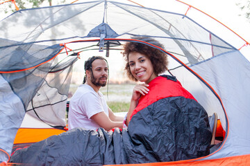 young african american couple on a hike lying in a tent in sleeping bags in the forest in nature, woman covering herself with a warm comfortable sleeping bag and smiling on a journey