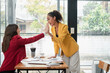 © Satori Studio - Two professional women in vibrant blazers high-fiving at a desk, celebrating success in a contemporary office environment.