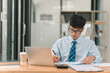 © phaitoon - A man wearing a blue tie is sitting at a desk with a laptop and a stack of papers. He is writing on the papers with a pen. The scene suggests a professional setting