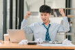 © phaitoon - A man in a blue shirt and tie is celebrating with his hands raised in the air. He is sitting at a desk with a laptop and a calculator