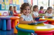 © Peeradontax - A joyful young girl sits at a vibrant, colorful table in a lively classroom, happily using a tablet.