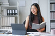 © amnaj - Smiling asian businesswoman analyzing financial data on digital tablet and taking notes on a book