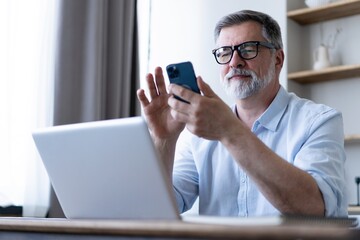 Sticker - Handsome smiling senior man wearing glasses using mobile phone while sitting at his workplace with laptop at home