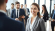 © halo - A young businesswoman stands in the foreground, smiling and looking at the camera while talking to other people standing around her in an office with large windows.