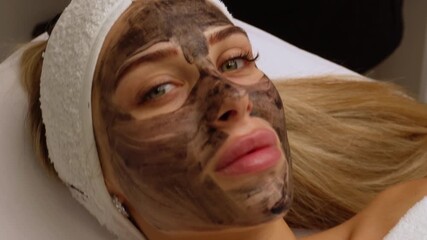 Wall Mural - cosmetologist applying black mask on pretty woman face wearing black gloves, gorgeous woman in spa having facial procedures