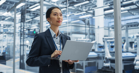 Thoughtful Asian Engineer Monitoring and Analyzing Conditions at a Pristine Modern Electronics Factory with Automated Robot Hands Working with Help of Artificial Intelligence Software