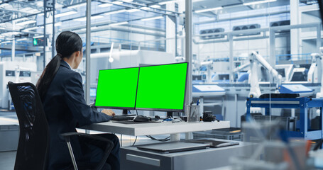 Fototapete - Female Robotics Technician Working on a Desktop Computer with Green Screen Chromakey Mock Up Display in a Factory Office. Technological Research and Development Facility with Autonomous Assembly Line